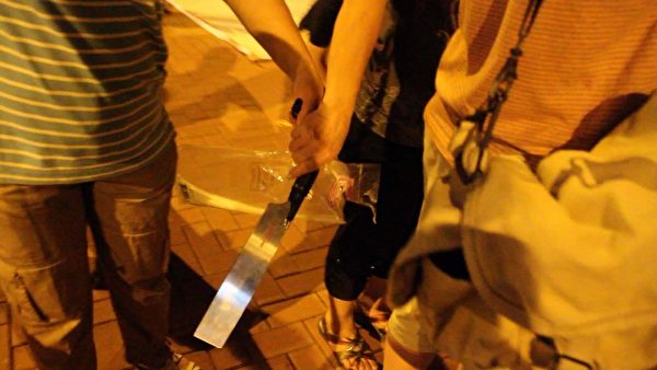 7月4日晚，「香港青年關愛協會」在中港邊境落馬洲出動鋸刀等械具，恐嚇在場的法輪功學員和採訪記者，而在場警員卻袖手旁觀不作為。圖為一名穿間條衫的女兇徒一度從另一名協會徒眾手上接過鋸刀，準備趨前恐嚇記者。（攝影：蔡雯文／大紀元）