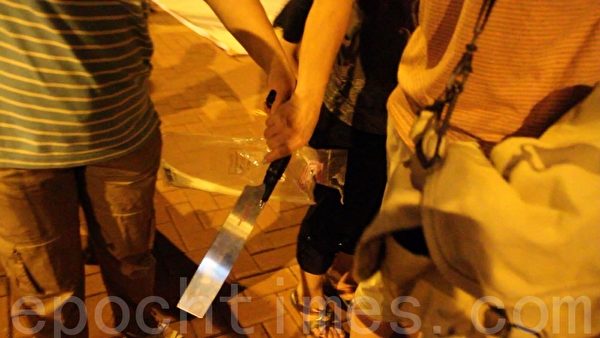 2012年7月4日晚，“香港青年关爱协会”在中港边境落马洲出动锯刀等械具，恐吓在场的法轮功学员和采访记者，而在场警员却袖手旁观不作为。图为一名穿间条衫的女凶徒一度从另一名协会徒众手上接过锯刀，准备趋前恐吓记者。（摄影：蔡雯文／大纪元）