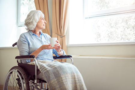 高年孤独的女人坐在轮椅上，手拿着茶看着窗外。